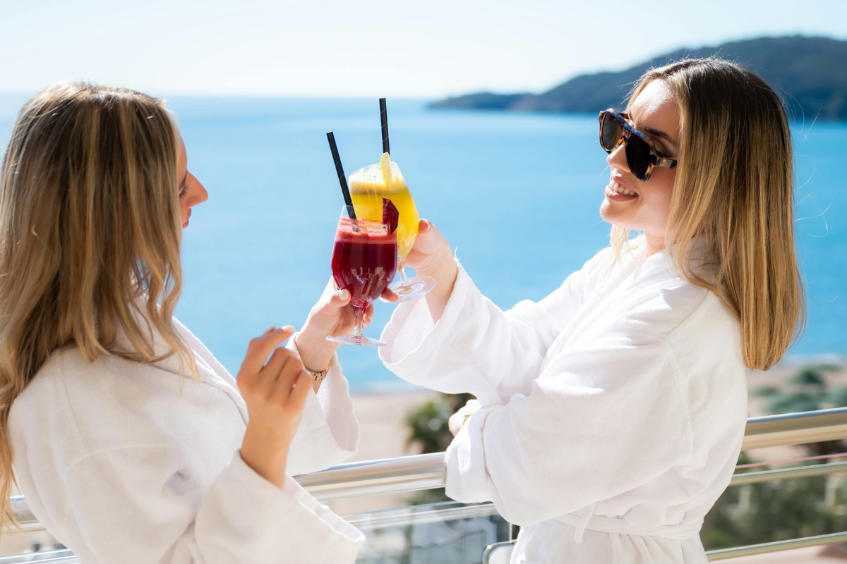 Two women in white robes stand on an ocean-view balcony, smiling and clinking together an orange cocktail, with a blue sea and hazy coastline in the background.