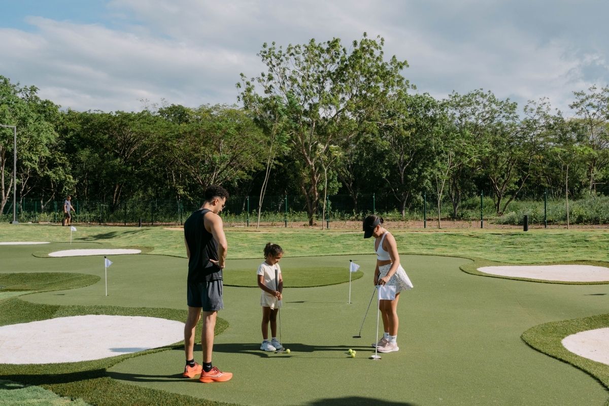 A man, woman, and child playing on a putting green at the One Hacienda Pinilla development with a forest background.