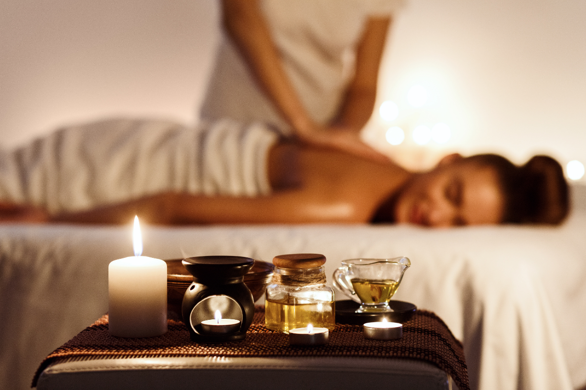 A woman relaxes face-down receiving a back massage in a softly lit spa setting, with a lit candle, massage stones, and small bottles of oil arranged on a tray in the foreground.