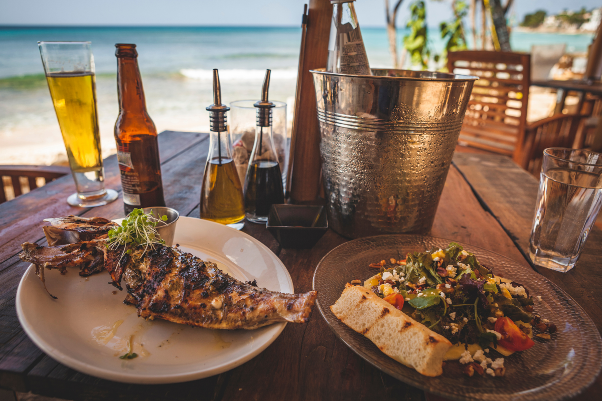 Grilled fish and salad served on an outdoor dining table near the ocean with drinks.