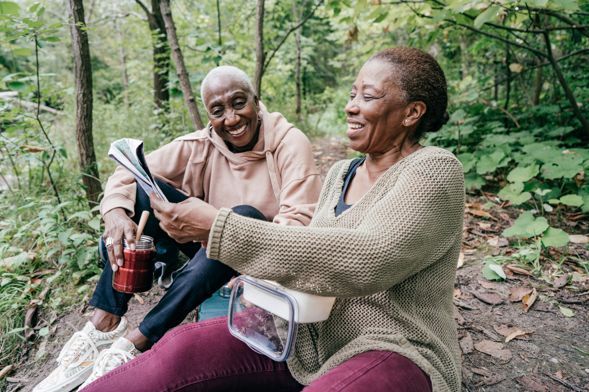 Two women sitting on a forest trail, laughing and looking at a map while enjoying a hike.