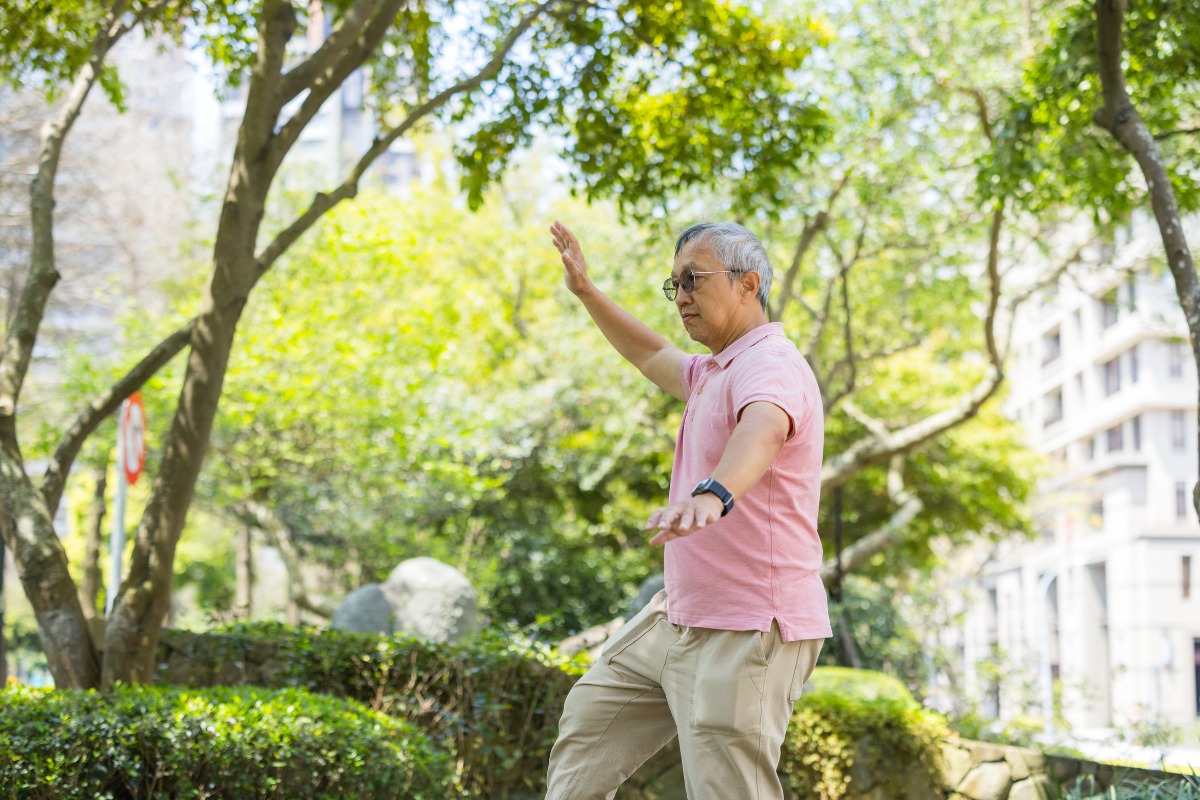 Older man balancing on a railing or low wall in a park, arms outstretched for stability, surrounded by trees.